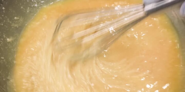 Close-up shot of a chef whisking a bright orange mixture of raw eggs or batter in a metal bowl using a wire whisk. The motion creates a smooth, swirling texture, capturing the essential preparation st