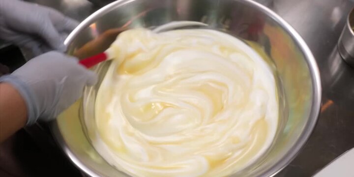 Close-up stock footage of a professional baker wearing blue nitirle gloves using a red spatula to gently fold a smooth egg yolk mixture into a fluffy, white whipped meringue batter. The preparation ta