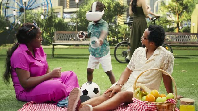 Boy wears a VR headset and raises both controllers while seated with his parents on a picnic blanket during a sunny family outing in an urban park, playing with virtual reality technology.
