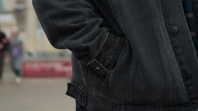 Close up of person wearing dark denim jacket with their hand hanging by side gradually moving to place it in front. subject clothing includes visible fabric texture and stitching details