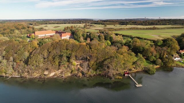 Drone footage of a historic manor estate surrounded by colorful autumn woodland and calm coastal water, with the Little Belt Bridge visible in the distance.