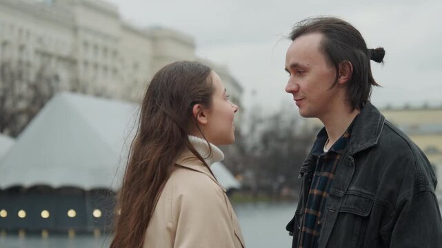 Close up of young woman and man standing outdoors in urban setting engaged conversation with soft lighting. Woman touches side of smiling boy face while they lean towards each other