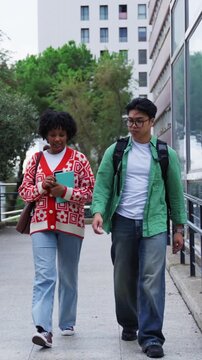 Vertical. Two university students walk down campus steps after class, chatting and smiling in a casual outdoor setting. Mixed ethnicity pair with bag and notebook, relaxed mood academic environment.