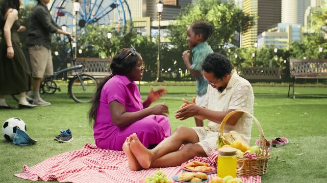 Young boy dances and has fun with his parents on a checkered blanket, fooling around and laughing together with silly moves during a picnic.