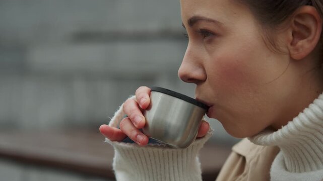 Close up of woman drinking from metal cup while wearing white turtleneck sweater. scene is set outdoors with soft lighting and urban background focusing on action of sipping beverage
