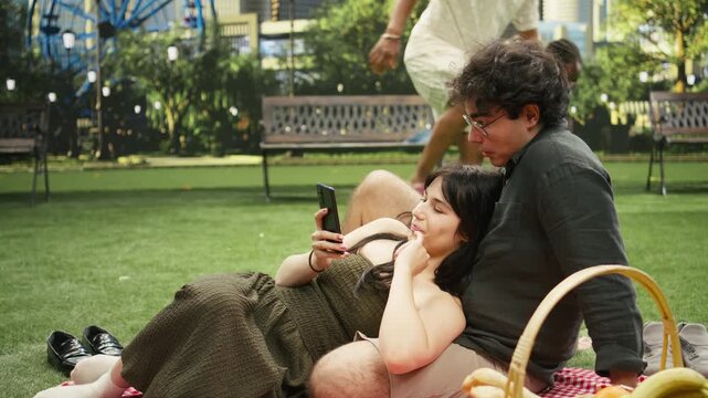 Boyfriend laughs at online reels with girlfriend leaning on him, resting on picnic blanket and making jokes together during outdoor date.