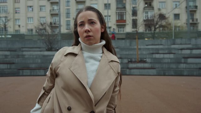 Close up of young woman performing arm movements in outdoor urban setting with buildings and stone steps visible. Woman wears beige trench coat over white turtleneck her long hair moving