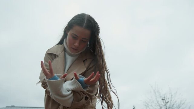 Close up of young woman performing expressive dance movements outdoors under overcast sky. Woman wears beige trench coat and white turtleneck with her long hair flowing as woman moves