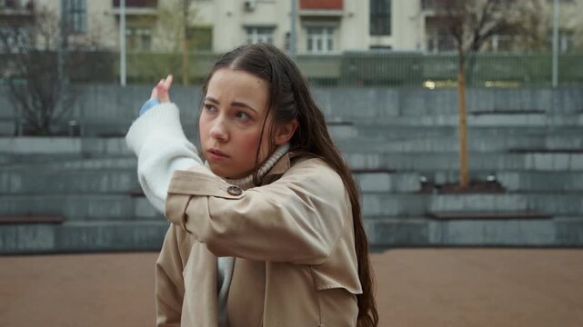 Close up of young woman performing martial arts movements outdoors in front of concrete steps and buildings. Woman wears beige trench coat over white turtleneck with visible red marks on her