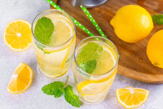 Refreshing homemade lemonade in glasses with fresh lemon slices, mint leaves, ice cubes, sparkling water served on wooden tray on white tiled kitchen background. Summer citrus beverage, drink concept.