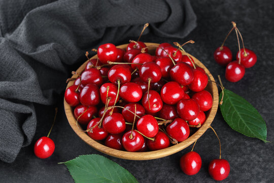 Fresh ripe red sweet cherries in wooden bowl on dark textured stone background with green leaves and black linen towel. Summer fruit, berry, harvest, healthy organic food