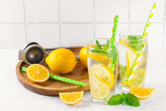 Refreshing homemade lemonade in glasses with fresh lemon slices, mint leaves, ice cubes, sparkling water served on wooden tray on white tiled kitchen background. Summer citrus beverage, drink concept.