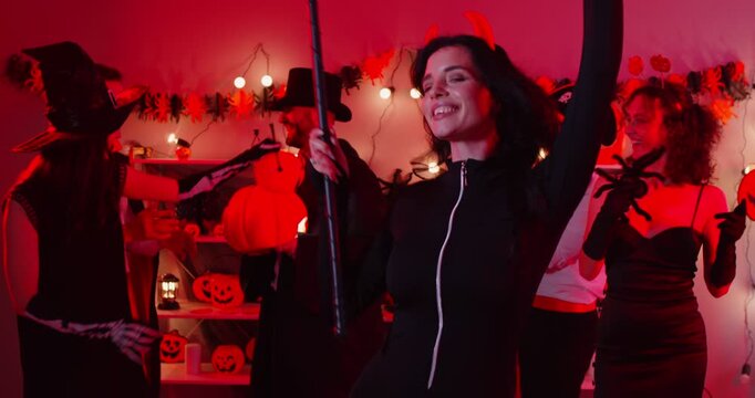 Young happy woman in black devil costume dancing with friends at Halloween party holding scythe in hands. People group having fun celebrating holiday at home. Celebration concept. 4k video.