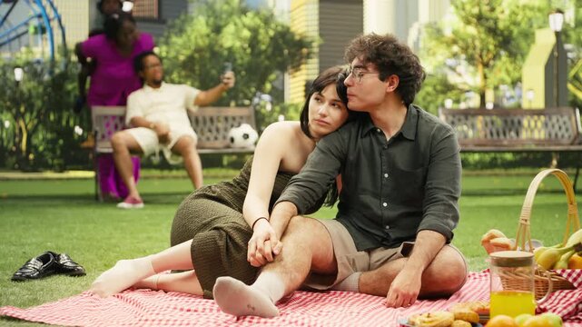 Romantic couple sit on a picnic blanket and caress each other while talking together during a relaxed outdoor date with an urban skyline in the background, showing affection during their picnic date.