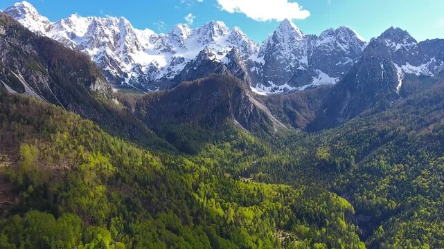 Aerial view of Triglav mountain range with snow-capped peaks above lush green forests under a clear blue sky in Mojstrana, Jesenice, Slovenia.