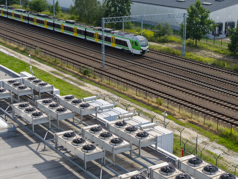 Aerial view of an industrial data center cooling system featuring rows of HVAC fans on a rooftop, situated beside a railway track with a passing commuter train in Poland.