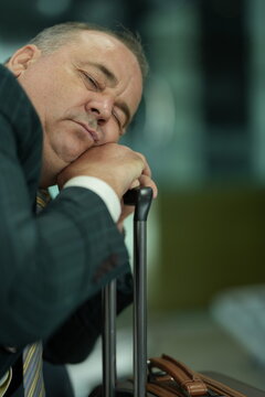 A tired male passenger falls asleep with his head on the suitcase handle on the seats of a departure gate, a familiar image of travel exhaustion, showing jet lag and long haul fatigue