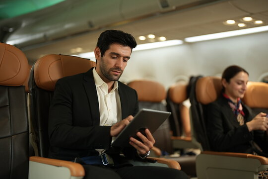 A male business traveller with a tablet reads a tablet in his aircraft seat on board a commercial flight, in the warm light of the aircraft cabin, with a flight attendant working nearby