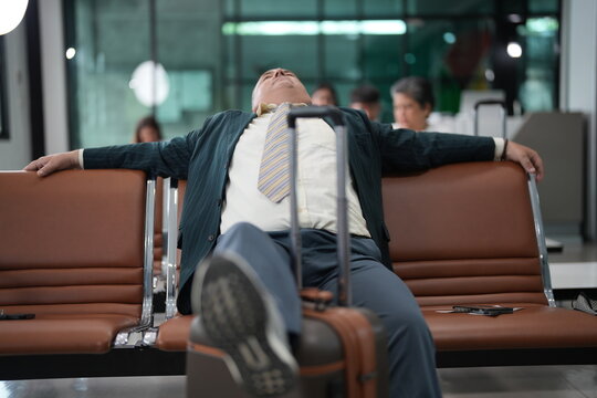 A senior businessman stretched across the seats sprawls across the bench during a long delay in a quiet part of the departure hall, with his suitcase standing beside him
