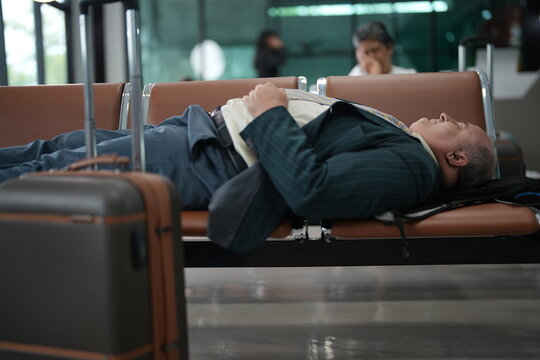 An exhausted traveller asleep on a bench sleeps on the seating row beside his suitcase in an airport departure lounge, showing what a long delay really looks like