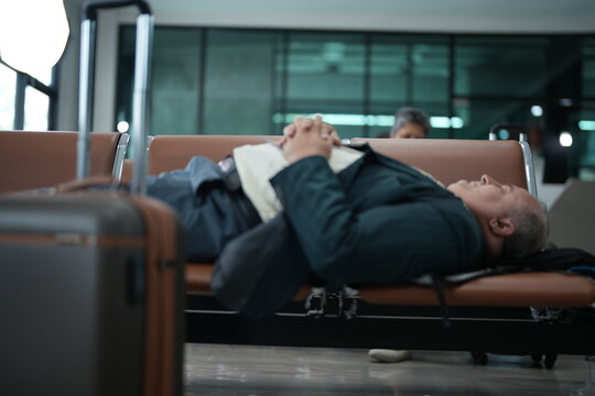 A weary traveller resting on the terminal seating dozes on the airport seats with his jacket on in a quiet corner of the departure hall, with the terminal glass glowing behind