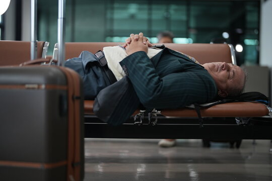 An exhausted traveller asleep on a bench stares at his screen from a horizontal position on the seating row of an airport gate, with the terminal glass glowing behind
