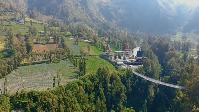 Aerial view of Jembatan Kaca glass bridge spanning a deep valley surrounded by lush green terraced fields and dense forest in Kecamatan Sukapura, Jawa Timur, Indonesia.
