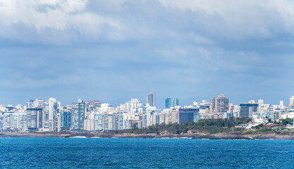 Naklejka premium Wide panorama of a modern coastal city skyline stretching along a rocky shore and deep blue sea under a soft, light blue sky filled with white fluffy clouds on a bright clear day