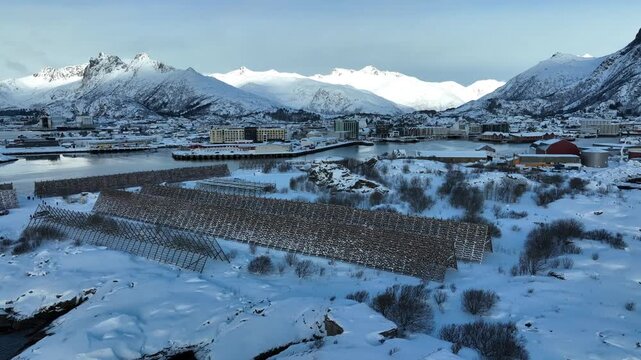 Aerial view of Svolvaer with traditional wooden fish drying racks set against snow-covered mountains and a frozen harbor in Svolvaer, Nordland, Norway.