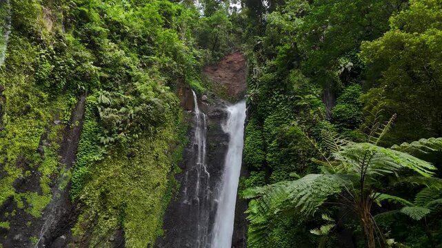 Aerial view of Curug Ciarjuna waterfall cascading down a lush tropical cliff surrounded by dense green forest foliage in Garut, West Java, Indonesia.