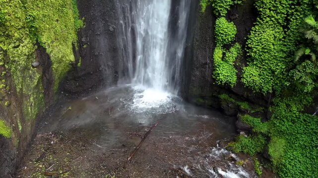 Aerial view of the cascading Curug Ciarjuna waterfall surrounded by lush green vegetation and mossy rock cliffs in Garut, West Java, Indonesia.