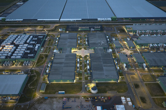 Aerial view of a large data center complex illuminated at night, featuring rows of server buildings, cooling infrastructure, and surrounding greenhouses in the Netherlands.