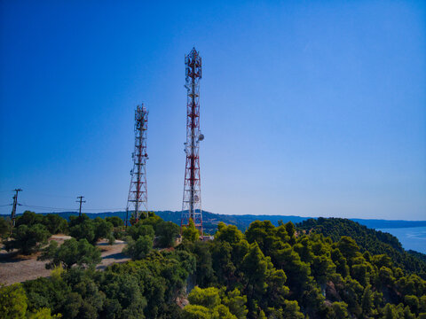 Aerial view of two tall telecommunication towers standing amidst lush green pine trees on a hill overlooking the sea in Possidi, Greece.