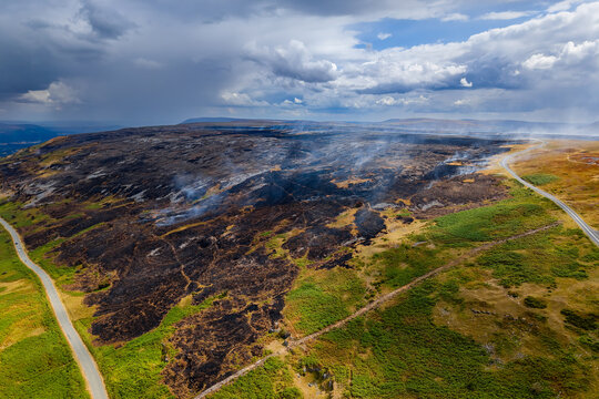 Scorched ground and rising smoke after wildfire on Llangynidr Moors, aerial view across charred upland landscape bounded by country roads, Brecon Beacons, Wales