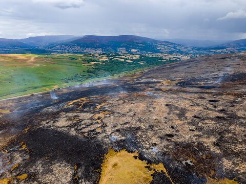 High angle aerial view of scorched ground after hill fire, contrast between burnt black heather and green farmland in valley below, wildfire destruction