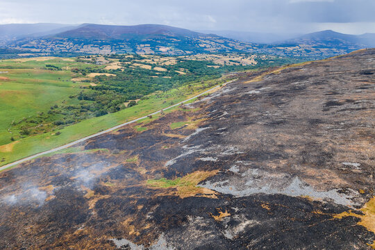 Elevated view of wildfire damage across upland heath, dark ash contrast with green farmland below, documenting environmental destruction from rural bushfire