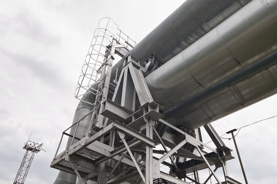 The photo shows a pipeline against a gray sky, and a tower with floodlights in the background