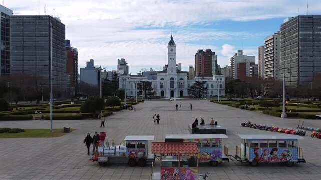 Dynamic drone approach over Plaza Moreno moving towards the city hall facade with people walking on a cloudy day in La Plata, Argentina.