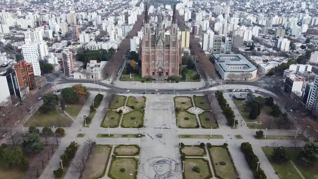 High altitude panoramic drone shot of Plaza Moreno featuring city traffic and La Plata Cathedral facade on a cloudy day, Argentina.