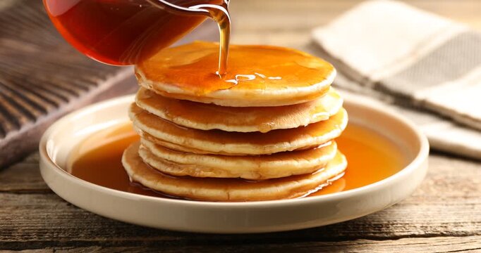 Pouring maple syrup onto pancakes at wooden table, closeup