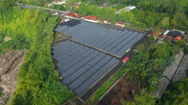 Aerial view of PLTS 1MWp Bangli solar power plant surrounded by lush green trees and rural landscape in Bangli, Bali, Indonesia.