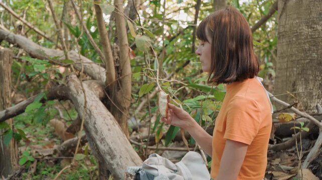 Close up of woman examining and eating piece of bread in forest setting with dense green foliage and tree trunks. Woman holds partially eaten food while seated on ground next to bag