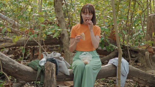 Close up of woman eating bread while sitting on log in forest. Woman holds and takes bites from pieces of food with bags containing more items placed beside her. scene features dense green