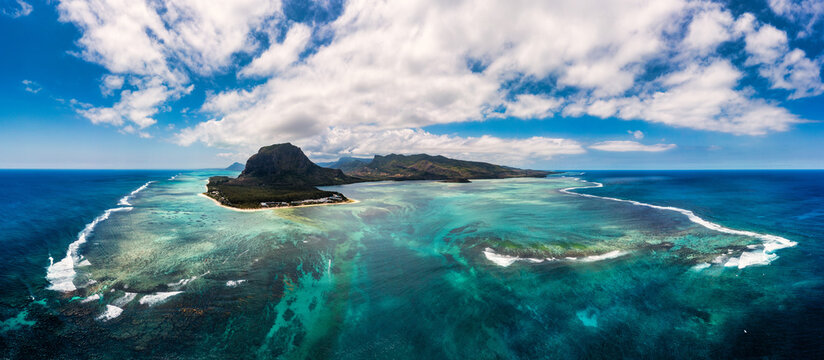Aerial view of Le Morne Brabant mountain peninsula surrounded by turquoise coral reef lagoons and deep blue ocean under a bright sky with scattered white clouds, Mauritius.