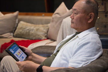 Fototapeta premium Senior Asian man sitting on cushioned chair at home holding tablet showing colorful app, copy space