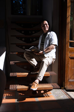 Senior Asian man sitting on floating wooden stairs near wooden framed glass door reading open book