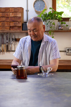 Senior Asian man sitting at kitchen counter in striped shirt holding phone, glass teapot visible