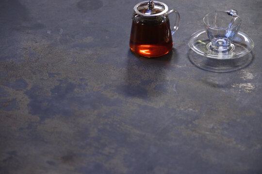 Clear glass teapot with metal lid and teacup on saucer resting on textured tabletop, copy space