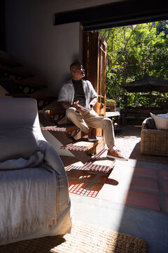 Senior Asian man sitting on wooden floating stairs at home reading book near open door
