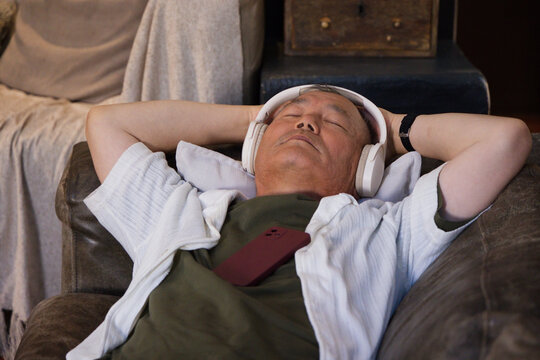 Senior Asian man reclining on sofa in living room, wearing headphones, phone on chest